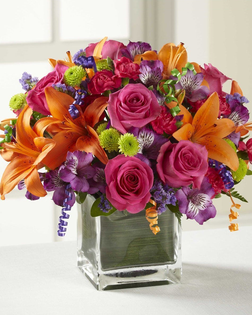 Celebrate! Standard (in a 5 x 5 inch Cube) A vibrant and festive bouquet of hot pink roses, orange lilies, purple alstroemerias and green button pops are accented with  ribbon and arranged in a leaf lined glass cube.  Approximate sizes:  Standard 9